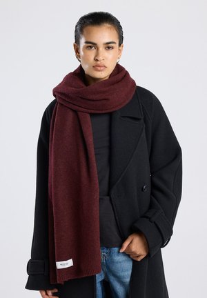 Bordeaux sjaal van zachte stof, om de nek gedrapeerd, in combinatie met een zwarte jas en jeans. Voorzien van een logolabel aan de onderrand.