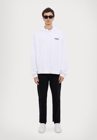 Hvit hettegenser med logo foran, kombinert med sorte bukser og hvite sneakers. Stoffet ser mykt ut og har en avslappet passform.