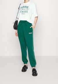 Vihreät urheiluhousut valkoisilla sivuviivoilla, yhdistettynä valkoiseen t-paitaan, jossa on vihreä teksti. Mustat kengät tekstuuripinnalla.