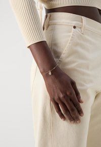 Pulsera de oro con un colgante en forma de lágrima. La mano descansa sobre una tela texturizada de color crema de unos pantalones de cintura alta.