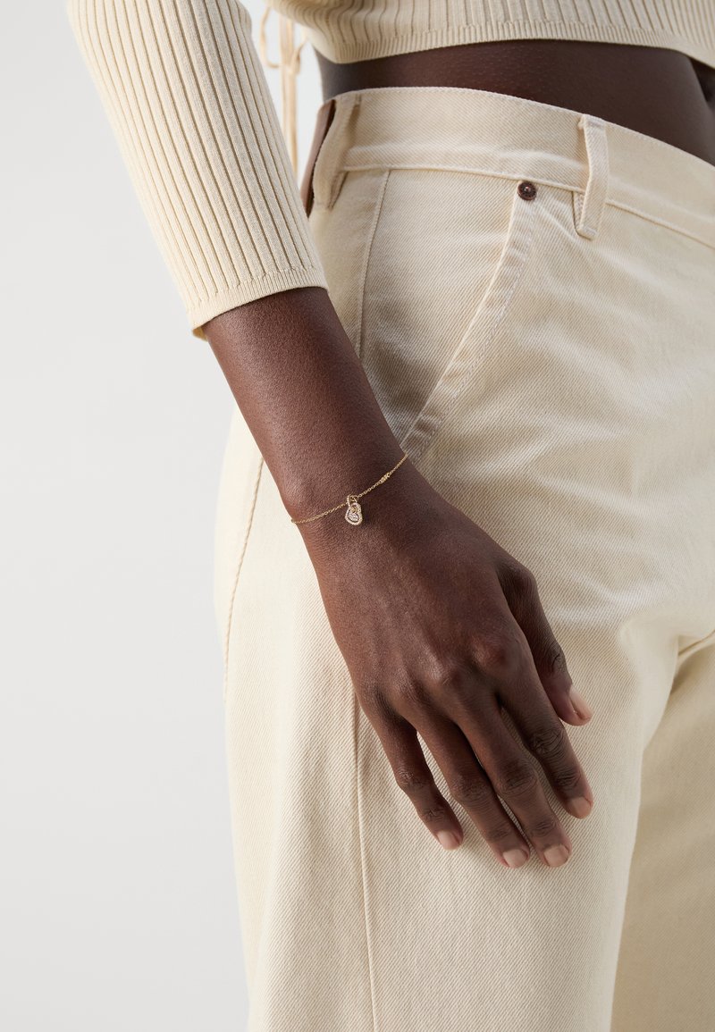 Pulsera de oro con un colgante en forma de lágrima. La mano descansa sobre una tela texturizada de color crema de unos pantalones de cintura alta.