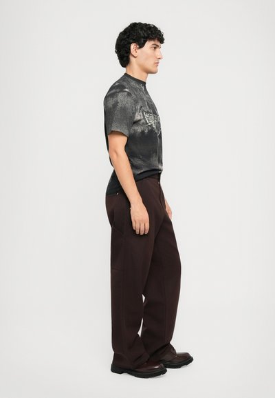 Man stands sideways wearing a black and gray graphic T-shirt, dark brown wide-leg pants, and dark brown shoes against a plain white background.