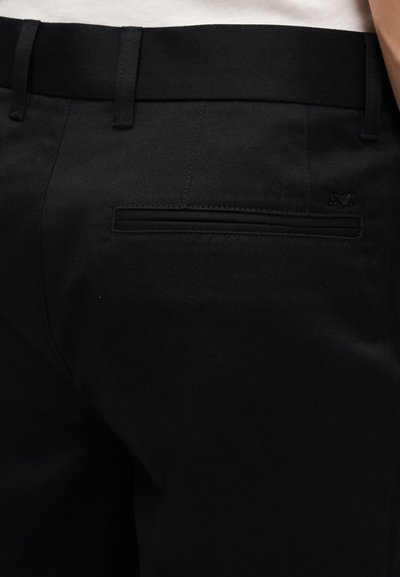 Κοντινό πλάνο πίσω τσέπης μαύρου παντελονιού με θηλιές για ζώνη και μικρό ανάγλυφο λογότυπο κοντά στην άκρη της τσέπης.