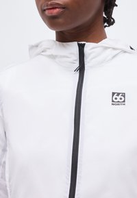 Gros plan d’une personne portant une veste à capuche blanche avec une fermeture éclair noire et un logo 66° NORTH sur la poitrine.