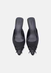 Μαύρα παπούτσια με μυτερή μύτη, στυλ slip-on, με μικρά φιόγκους στην κορυφή και την ένδειξη "HUGO" χαραγμένη στις εσωτερικές σόλες, τοποθετημένα σε λευκό φόντο.