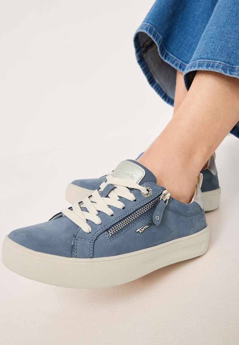 Baskets en daim bleu avec lacets blancs, fermeture éclair latérale et détail du logo, portées avec un jean en denim bleu, ourlé au-dessus des chevilles.
