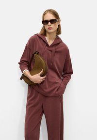 Burgundy hooded sweatshirt with a relaxed fit, paired with matching pants. Model holds a brown curved shoulder bag. Textured fabric.