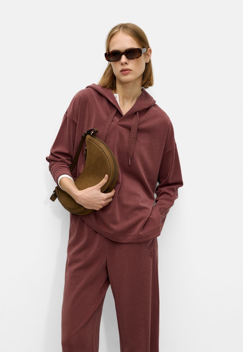 Burgundy hooded sweatshirt with a relaxed fit, paired with matching pants. Model holds a brown curved shoulder bag. Textured fabric.