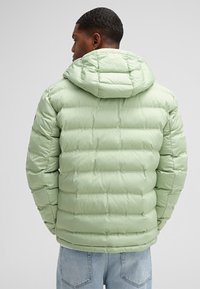 Giacca a vento verde chiaro con cappuccio, caratterizzata da sezioni trapuntate e una texture liscia. Indossata con jeans azzurri, vista da dietro.