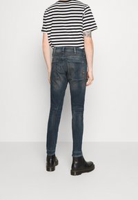 Person som bär mörkblå snäva jeans, svarta läderstövlar och en svartvit randig kortärmad skjorta, står med ryggen vänd.