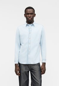 Camisa de ganga azul clara com mangas compridas, colarinho com botões e bainha reta, com botões cor de pele e textura suave. Combinada com jeans escuros.