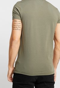 Zielona koszulka z krótkim rękawem, wykonana z miękkiej bawełny. Charakteryzuje się dopasowanym krojem, okrągłym dekoltem i prostym dołem. Widoczny tatuaż na ramieniu.