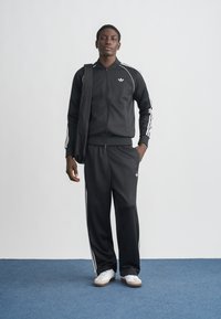 Vyras juodu Adidas sportiniu kostiumu su baltais dryžiais, baltomis sportinėmis avalynėmis, laikantis juodą krepšio diržą ant vieno peties, stovintis ant mėlyno kilimo.