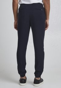 Man in een marineblauwe joggerbroek met elastische boorden en zwarte sneakers, staand met zijn rug naar de camera tegen een eenvoudige lichte achtergrond.