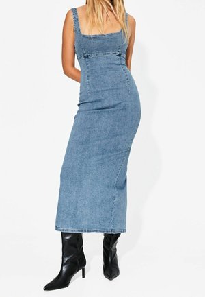 Femme portant une longue robe en jean bleu ajustée avec de larges bretelles et des bottes noires à talons pointus, debout sur un fond blanc.