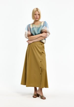 Frau mit kurzen blonden Haaren, die eine transparente, mehrfarbige Bluse mit Puffärmeln und einen langen senffarbenen Rock mit seitlichen Knöpfen sowie Sandalen mit Absatz trägt.