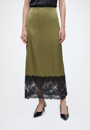 Woman wearing an olive green midi skirt with black lace hem, paired with black pointed-toe slingback heels and a black top.