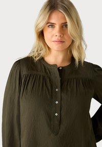 Blouse vert olive foncé avec un col rond, des détails plissés aux épaules et une fermeture à boutons avec de petits boutons en argent. Tissu doux et texturé.