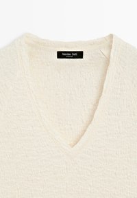 Bluzka z kremowego materiału o fakturze, z dekoltem w kształcie litery V i czarną metką Massimo Dutti przyszytą z tyłu wewnątrz kołnierza.
