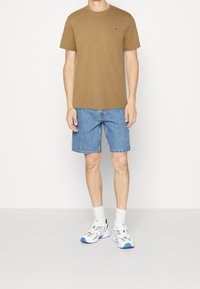 Brązowa bawełniana koszulka, niebieskie dżinsowe szorty, białe i niebieskie sneakersy oraz białe skarpetki. Minimalistyczny design z małym logo na koszulce.