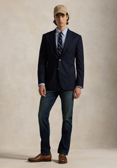 Tânăr purtând un sacou bleumarin, cămașă albastru deschis, cravată în dungi, blugi închiși la culoare, pantofi maro și o șapcă bej de baseball, stând în picioare pe un fundal neutru.