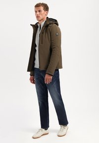 Olijfgroene jas met capuchon, rits aan de voorkant en zwarte accenten. Draag het over een lichtgrijze top, gecombineerd met donkere jeans en witte sneakers.