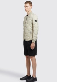 Beige quiltad puffjacka med vågmönster, dragkedja fram, ribbade ärmslut, bärs över svarta shorts och svarta sneakers. Minimalistisk design.