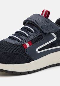 Sneaker Geox blu navy realizzato in materiale mesh e suede, con dettagli bianchi e rossi, cinturino regolabile e suola in gomma testurizzata per una migliore aderenza.