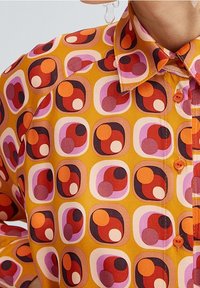 Yellow button-up shirt featuring a geometric pattern of circles in red, pink, and white. Textured fabric with a pointed collar and button details.