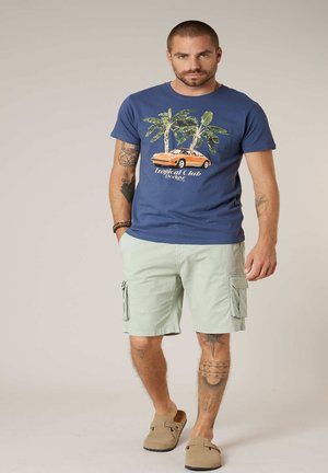 Homme tatoué portant un t-shirt à imprimé tropical bleu, un short cargo clair et des chaussures beige à enfiler, se tenant devant un fond uni.
