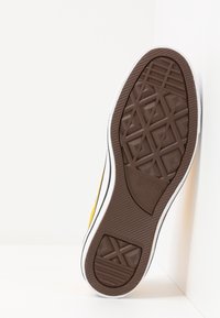 Semelle de chaussure en caoutchouc marron avec un motif texturé, bord blanc contrastant et dessus jaune visible sur le côté.