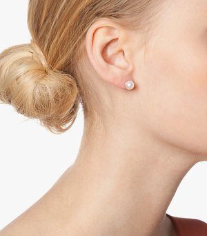 Round pearl stud earrings placed in a pierced ear, showcasing a lustrous, smooth surface. Simple design, light in color against skin tone.