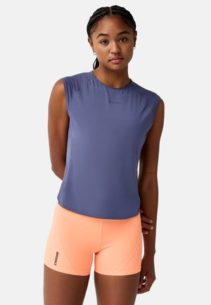 Jonge vrouw met gevlochten haar draagt een mouwloos paars topje en feloranje sportshorts tegen een effen witte achtergrond.
