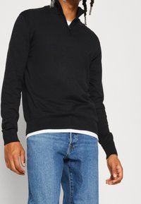 Schwarzer Zip-Hals-Pullover aus weichem Material mit langen Ärmeln und gerippten Bündchen, kombiniert mit hellblauen Jeans.