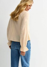 Beige Bluse mit langen Ärmeln aus strukturiertem Stoff, bestickten Blumenakzenten an den Ärmeln und Rüschensäumen, getragen mit blauen Jeans.