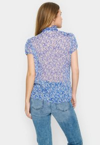 Bluză florală semi-transparentă de culoare albastră, cu mâneci scurte puff, guler înalt și un design stratificat cu modele albastre deschise și închise.