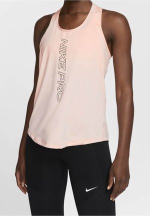 Mujer vestida con una camiseta sin mangas Nike Pro de color rosa claro y leggings negros de Nike, de pie frente a un fondo liso.