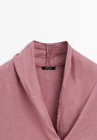 Dettaglio del colletto di una camicia di lino malva con scollo a V profondo e etichetta del marchio visibile cucita all'interno del collo.