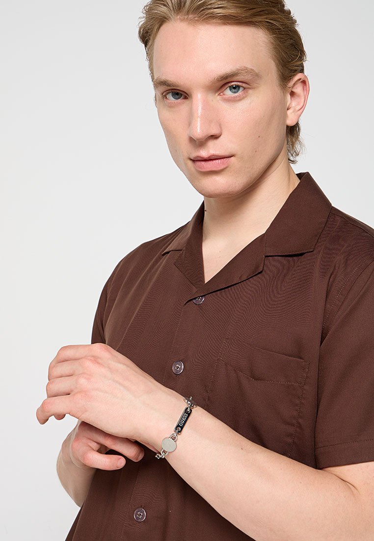 Chemise marron à manches courtes avec col, en tissu texturé, et boutons violets. Un bracelet en argent avec des accents ronds et rectangulaires est porté.