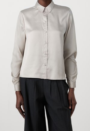 Osoba ubrana w jasnoszarą satynową bluzkę na guziki z długim rękawem oraz czarne, luźne spodnie, widoczna od ramion do bioder.