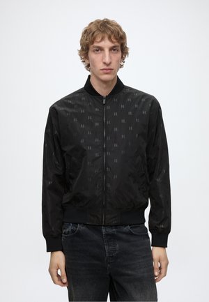Jeune homme aux cheveux blonds bouclés portant une veste noire zippée avec un motif subtil et un jean foncé, debout devant un fond blanc uni.