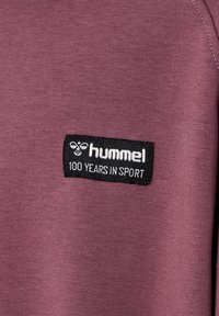 Étiquette noire avec logo hummel blanc et texte "100 ans dans le sport" sur un tissu violet d'un vêtement.