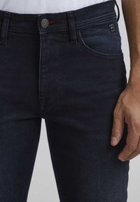 Jeans de mezclilla azul oscuro con corte ajustado, que cuentan con bolsillos frontales, un botón de metal y sutiles detalles de costura en la cintura.