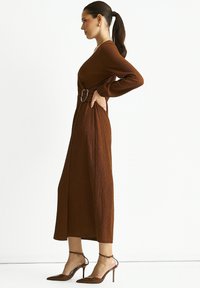 Brown textured midi dress with long sleeves and a belt featuring a silver buckle. Worn with transparent heeled shoes.