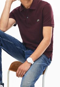 Burgundská polo košile s krátkým rukávem, dvoubotkovou légou a logem Lacoste. Oblečená s modrými džíny a bílými náramkovými hodinkami, sedící na židli.