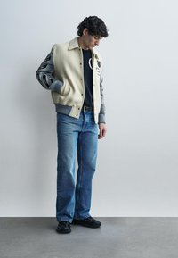 Νέος άνδρας που φοράει κρεμ και γκρι varsity μπουφάν, μαύρο πουλόβερ, μπλε τζιν και μαύρα παπούτσια, στέκεται μπροστά σε απλό ανοιχτό γκρι τοίχο.