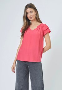 Blusa a maniche corte, con scollatura a V di un vivace rosa, realizzata in tessuto morbido, con dettagli arricciati sulle spalle, abbinata a pantaloni grigi a gamba larga.