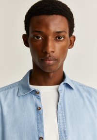 Lyseblå denimskjorte med knapkrave, prydet med brune knapper og en hvid t-shirt med rund hals under. Glat tekstur.