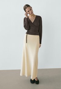 Brauner Wickelpullover mit langen Ärmeln, kombiniert mit einem cremefarbenen Maxirock. Der Rock hat eine weiche Textur, und schwarze Ballerinas runden das Outfit ab.