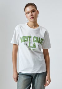 Hvid bomulds t-shirt med "WEST COAST" i grøn tryk, kortærmet, rund hals, afslappet pasform, og bæres med lyseblå denimjeans.